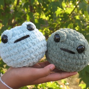 May include: Two crocheted frog plushies, one light blue and one dark green, with black eyes and a black mouth. The plushies are held in a person's hand.