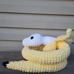 May include: A handmade crochet snake toy in shades of yellow and white. The snake's head is white with a black eye, and the body is coiled in a yellow hue. The tail has a black tip. The toy is made of soft, textured yarn.