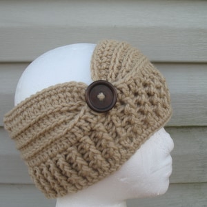 Puede incluir: Una diadema de ganchillo color beige con un diseño acanalado y un botón grande de color marrón oscuro. La diadema está envuelta alrededor de una cabeza de maniquí blanca, mostrando su textura y estilo. El color neutro la hace versátil.