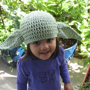 May include: A light green crocheted hat with large, pointed ears. The hat is worn by a young child wearing a purple long-sleeved shirt. The background features green plants and a blue pot.