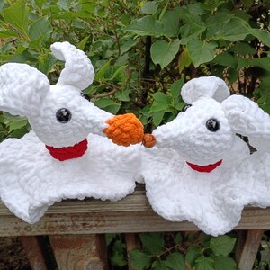 May include: Two white crocheted ghost dog figures with floppy ears, black button eyes, and red collars. One figure has a textured orange nose, while the other has a small brown nose. The figures are positioned facing each other, appearing to touch noses.