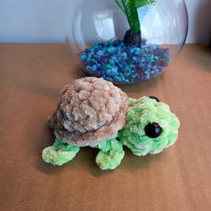 May include: A handmade plush turtle with a light brown shell and green body. The turtle has large black eyes and is made of soft, textured yarn. A glass fishbowl with blue and green stones and a green plant is in the background.