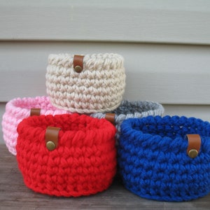 May include: Five crocheted baskets in different colors, including red, pink, white, gray, and blue. Each basket has a brown leather handle with a brass button.