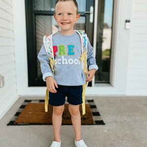 Puede incluir: Un niño pequeño con una sudadera gris con las palabras "PRE school" en letras de colores. Lleva pantalones cortos azules y una mochila amarilla con estampado floral. Está de pie sobre una alfombra de bienvenida con un patrón de cuadros negros y blancos.