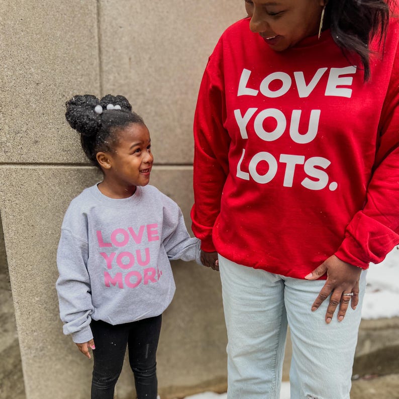 May include: A mother and daughter wearing matching gray and red sweatshirts with the text "Love You" and "Love You More" in pink. The mother's sweatshirt is red with white text and the daughter's sweatshirt is gray with pink text.