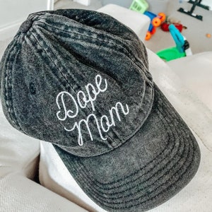 May include: A black denim baseball cap with white embroidered text that reads "Dope Mom".