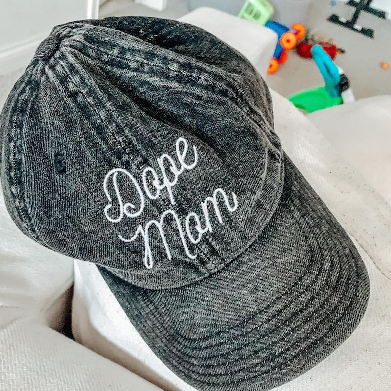 May include: A black denim baseball cap with white embroidered text that reads "Dope Mom".