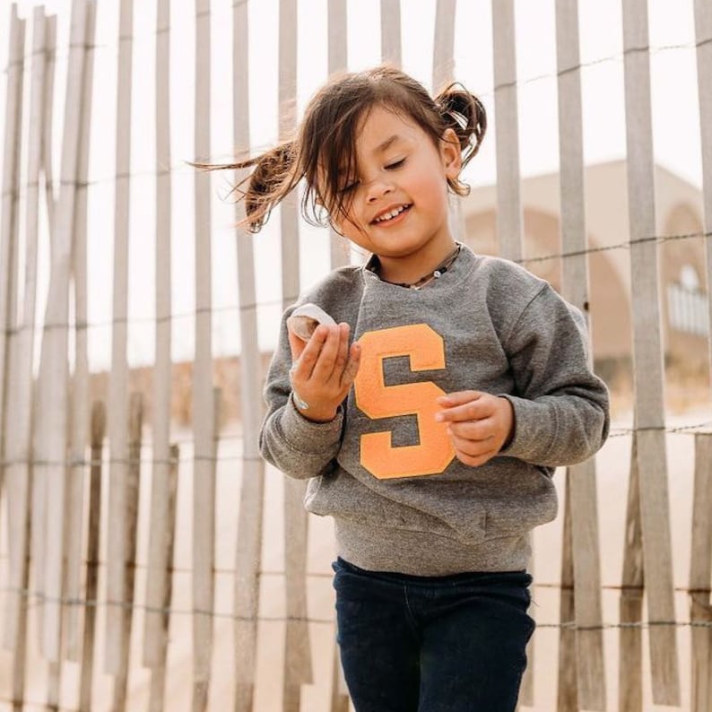 May include: A young girl wearing a gray sweatshirt with a large orange letter "S" on the front. She is holding a small white object in her hand and smiling.