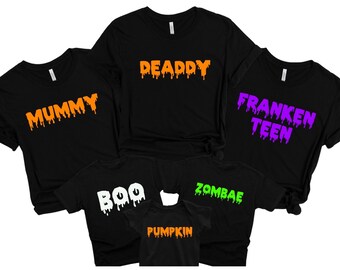 May include: Black t-shirts with Halloween-themed text in various colours. The shirts display the words "MUMMY," "DEADDY," "FRANKEN TEEN," "BOO," "ZOMBAE," and "PUMPKIN" in orange, white, green, and purple dripping font.
