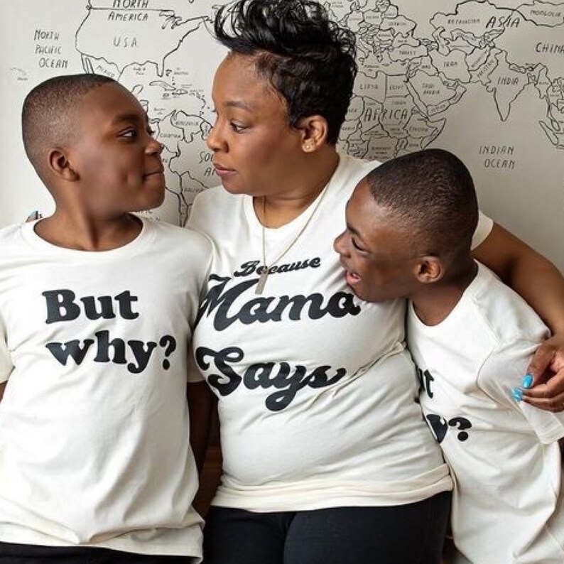 May include: A woman and two children wearing white t-shirts with black text. The woman's shirt says "Because Mama Says". The children's shirts say "But why?" and "But why?" The woman is standing in the middle with her arms around the children.