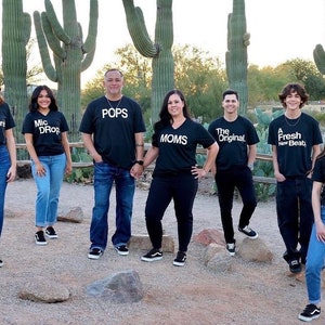 Peut inclure: Une famille de sept personnes portant des t-shirts noirs avec du texte blanc. Le texte sur les t-shirts est "The Encore", "Mic Drop", "Pops", "Moms", "The Original", "A Fresh New Beat", et "The Remix".