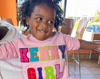 May include: A young child wearing a pink sweatshirt with the text "KENNY GIRL" in colorful, embroidered letters. The child has curly hair and a bow in their hair.