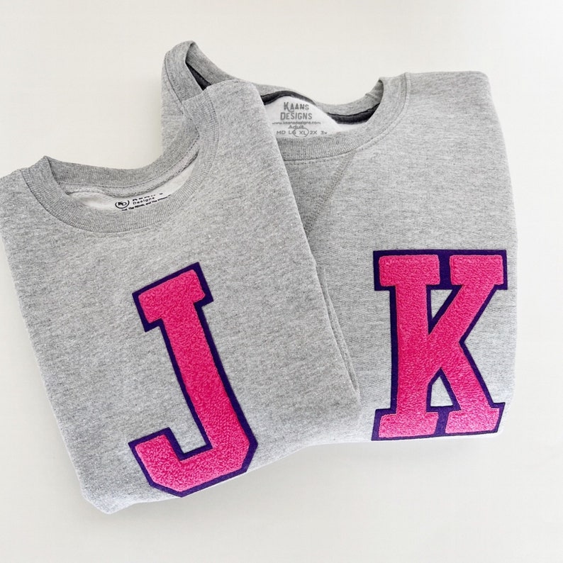 May include: Two gray sweatshirts with large pink felt letters, one with a "J" and the other with a "K". The letters have a dark purple outline.