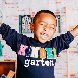 Puede incluir: Un niño pequeño con una sudadera azul marino con las palabras "kindergarten" en letras de colores. Está sonriendo y tiene los brazos extendidos.