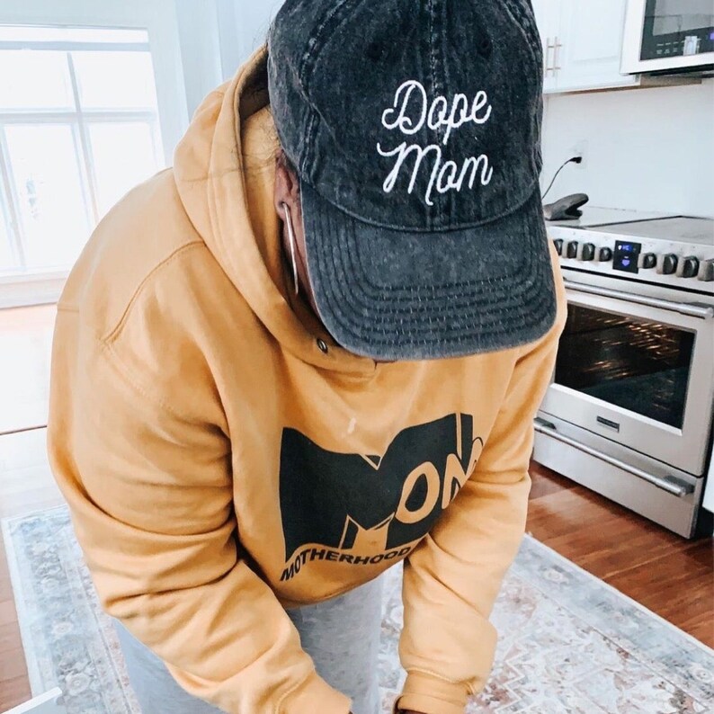 May include: A person wearing a mustard yellow hoodie with a black logo that says "Motherhood" and a black denim baseball cap with white embroidery that says "Dope Mom".
