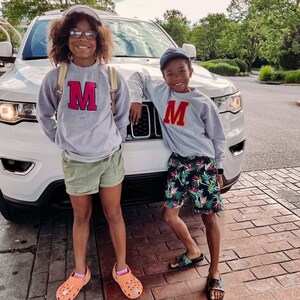 May include: Two children, a girl and a boy, stand in front of a white SUV. The girl is wearing a gray sweatshirt with a red "M" on the front, green shorts, and pink and orange sandals. The boy is wearing a gray sweatshirt with a red "M" on the front, blue and green floral print shorts, and black sandals.