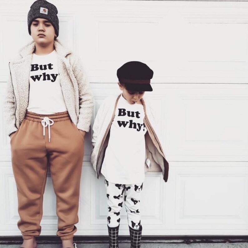 May include: Two children wearing white t-shirts with the text "But why?" printed on them. The child on the left is wearing a brown jacket and brown pants. The child on the right is wearing a black hat and black and white patterned pants.