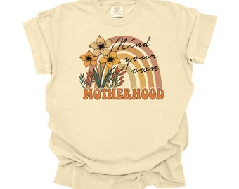 Puede incluir: Una camiseta beige claro con un arcoíris y un diseño floral. El texto "Mind your own Motherhood" está impreso en la camiseta.