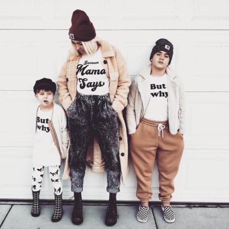 May include: A family of three wearing white t-shirts with black text. The adult is wearing a brown coat, black jeans, and a burgundy beanie. The two children are wearing brown jackets and beanies. The text on the shirts reads "Because Mama Says", "But Why", and "But Why".