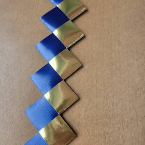 May include: A zig-zag pattern of alternating blue and gold diamond-shaped ribbon pieces. The ribbon is arranged diagonally against a light brown wooden surface. The gold sections have a metallic sheen.
