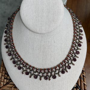 May include: A beaded necklace with a dark red and bronze color scheme. The necklace is displayed on a white mannequin bust. The intricate design features small, round beads and decorative floral-like clusters.