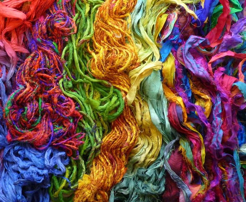 May include: A colorful assortment of silk ribbon scraps in shades of red, orange, yellow, green, blue, purple, and pink. The ribbons are tangled and overlapping, creating a vibrant and textured background.