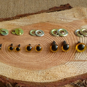 May include: Assortment of plastic safety eyes in various sizes with black pupils and amber colored irises. The eyes are on a wooden surface with metal washers for attaching the eyes.