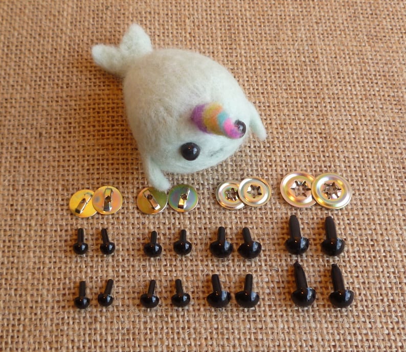 以下が含まれることがあります： クラフト用の黒い安全ピンアイが18個セットになっています。6組のアイと6セットの金属製ワッシャーが付いています。アイは茶色のジュート生地の背景に並べてあります。虹色の角を持つ小さな白いフェルトのナルトゥクも写っています。
