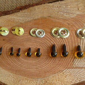 May include: Assortment of plastic safety eyes in various sizes, with black pupils and amber colored irises. The eyes are attached to black plastic posts. Also included are metal fasteners for attaching the eyes to fabric.