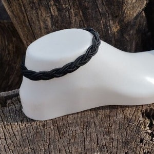 Può includere: Un braccialetto alla caviglia intrecciato nero su un piede di manichino bianco. Il piede è appoggiato su un ceppo d'albero marrone e ruvido.