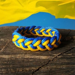 May include: Braided bracelet in blue and yellow, resting on a wooden surface. The bracelet's colors mirror the Ukrainian flag waving in the background. The bracelet is a symbol of solidarity and support.