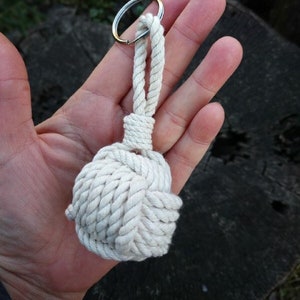 Op de afbeelding: Een witte touw sleutelhanger met een geknoopte bal aan het einde. De sleutelhanger is bevestigd aan een zilveren sleutelring.