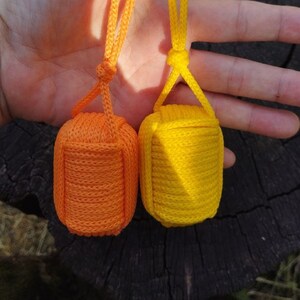 以下が含まれることがあります： 2つの装飾的なモンキーフィストキーホルダー。1つは明るいオレンジ色、もう1つは黄色です。各キーホルダーは、キーリングに取り付けるためのループが付いた、しっかりと織られた球形の結び目です。キーホルダーは、暗く質感のある背景に表示されています。