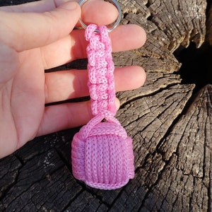 May include: A pink macrame keychain with a silver key ring. The keychain features a woven design and a small, rounded, knitted bottom. The item is held against a weathered, dark wood background.