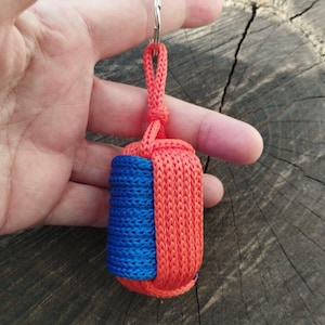 May include: A handmade keychain featuring a cylindrical design with a vibrant color scheme. The keychain is primarily orange with a blue accent, and includes a silver-colored keyring. The item is made of woven cord.