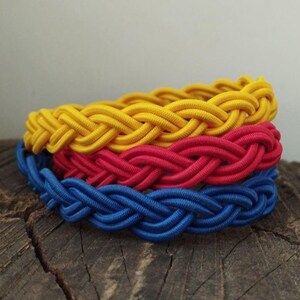 May include: Three braided bracelets in yellow, red and blue colours. The bracelets are stacked on top of each other.
