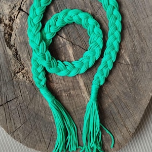 May include: Bright green braided rope with tassels. The rope is arranged in a circular pattern with two tassels at the ends. The rope is made of a soft, woven material. The background is a wooden surface.