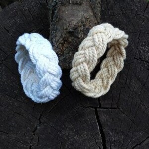 May include: Two braided rope bracelets, one white and one beige, on a dark brown wooden surface.