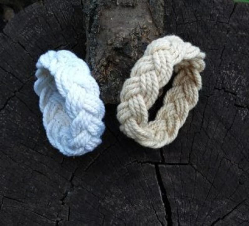 May include: Two braided rope bracelets, one white and one beige, on a dark brown wooden surface.