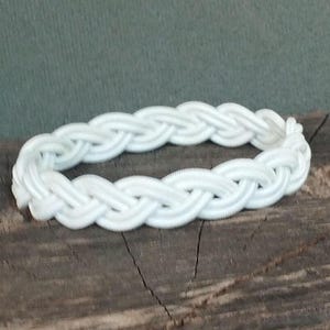May include: A white braided bracelet. The bracelet is made of thick, woven material, creating a textured appearance. The bright white color contrasts against the dark wooden surface it rests upon. The bracelet is a complete circle.