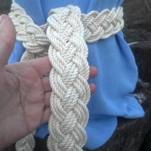 May include: Braided cream-coloured rope belt. The belt is thick and appears to be made of tightly woven rope. The belt is shown against a light blue background.