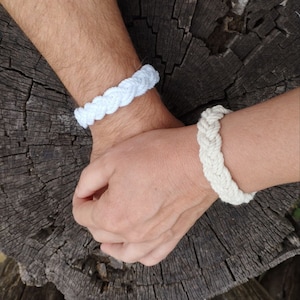 Könnte beinhalten: Zwei geflochtene Armbänder, eines weiß und eines cremefarben, werden gezeigt. Die Armbänder bestehen aus einem dicken, gewebten Material. Die Armbänder werden auf einer dunklen, strukturierten Holzoberfläche gezeigt.