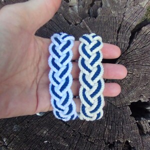 Op de afbeelding: Twee gevlochten touwarmbanden. De ene is wit met blauwe accenten, de andere blauw met witte details. De armbanden worden in een hand gehouden tegen een verweerde houten achtergrond.