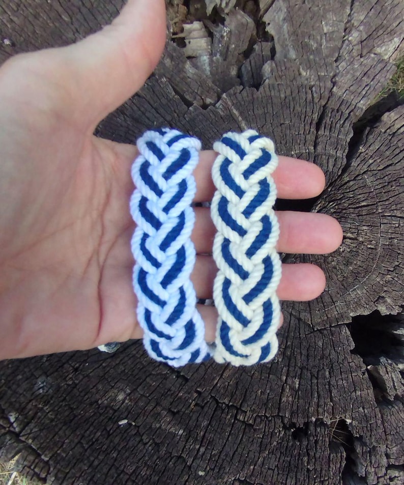 Op de afbeelding: Twee gevlochten touwarmbanden. De ene is wit met blauwe accenten, de andere blauw met witte details. De armbanden worden in een hand gehouden tegen een verweerde houten achtergrond.