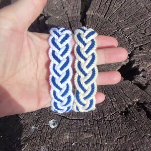 Op de afbeelding: Twee gevlochten armbanden, &eacute;&eacute;n wit met blauwe accenten en de andere gebroken wit met blauwe accenten, worden in een hand gehouden. De armbanden zijn gemaakt van dik, geweven touw. De achtergrond is een verweerde, bruine boomstronk.