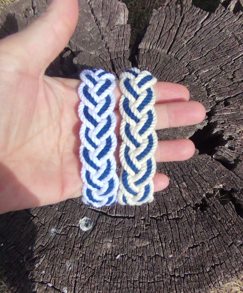 Op de afbeelding: Twee gevlochten armbanden, &eacute;&eacute;n wit met blauwe accenten en de andere gebroken wit met blauwe accenten, worden in een hand gehouden. De armbanden zijn gemaakt van dik, geweven touw. De achtergrond is een verweerde, bruine boomstronk.