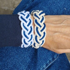 Op de afbeelding: Twee gevlochten touwarmbanden. E&eacute;n is wit met blauwe accenten, de andere is blauw met witte accenten. De armbanden zijn dik en lijken handgemaakt. De armbanden zitten om een pols, met een donkerblauwe mouw en een spijkerbroek op de achtergrond.
