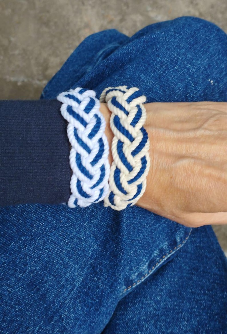 Op de afbeelding: Twee gevlochten touwarmbanden. E&eacute;n is wit met blauwe accenten, de andere is blauw met witte accenten. De armbanden zijn dik en lijken handgemaakt. De armbanden zitten om een pols, met een donkerblauwe mouw en een spijkerbroek op de achtergrond.