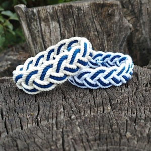 Op de afbeelding: Twee nautische armbanden van gevlochten touw in wit en marineblauw. De armbanden liggen op een verweerd, getextureerd houten oppervlak, wat een rustiek of maritiem thema suggereert.