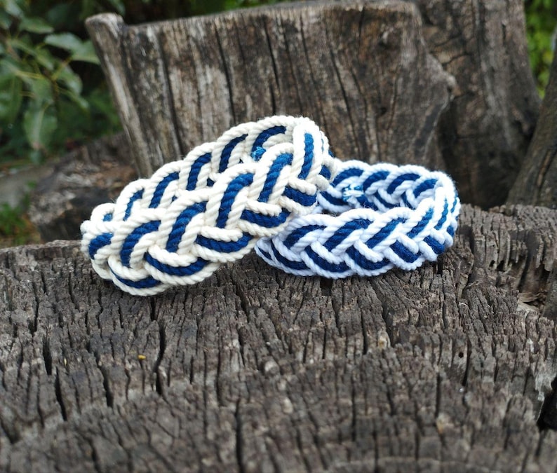 Op de afbeelding: Twee nautische armbanden van gevlochten touw in wit en marineblauw. De armbanden liggen op een verweerd, getextureerd houten oppervlak, wat een rustiek of maritiem thema suggereert.